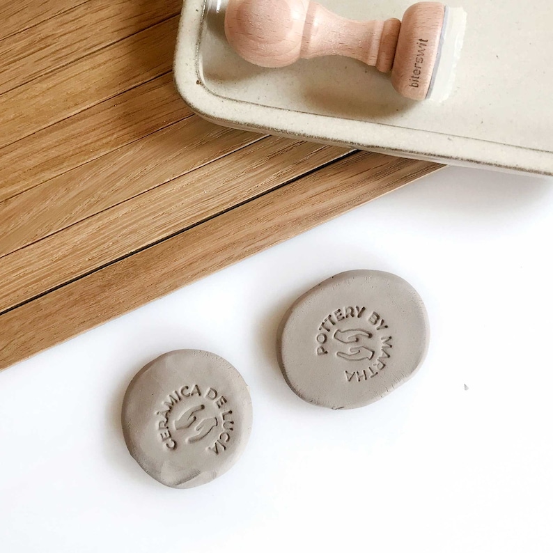 May include: Two gray ceramic stamps with the text "Ceramica de Lucia" and "Pottery by Martha" in a circular design. The stamps are on a white surface.