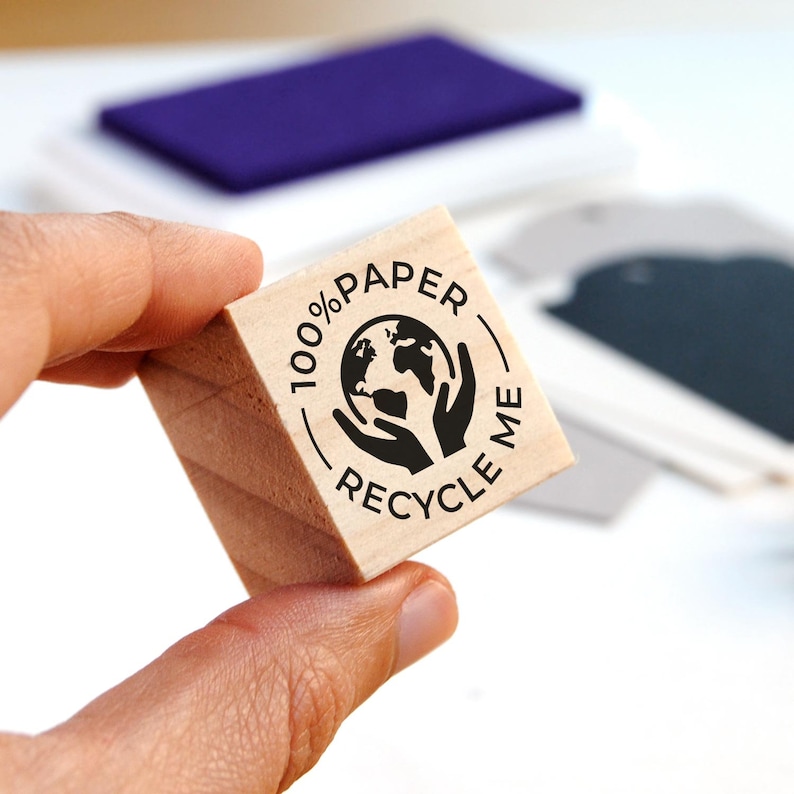 A hand holds the S-size 100% Paper Recycle rubber stamp, showing the detailed engraving on the wooden block. The compact design makes it easy to use on envelopes, small shipping boxes, and recyclable kraft paper. This eco-friendly branding tool is ideal for businesses looking to promote sustainable packaging and encourage recycling in a simple, effective way.