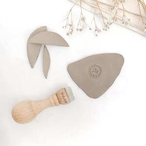 Puede incluir: Un sello de madera con el texto "Dierkswil" en &eacute;l. El sello est&aacute; al lado de un tri&aacute;ngulo de arcilla marr&oacute;n claro con un dise&ntilde;o circular que dice "Made by Pottery".