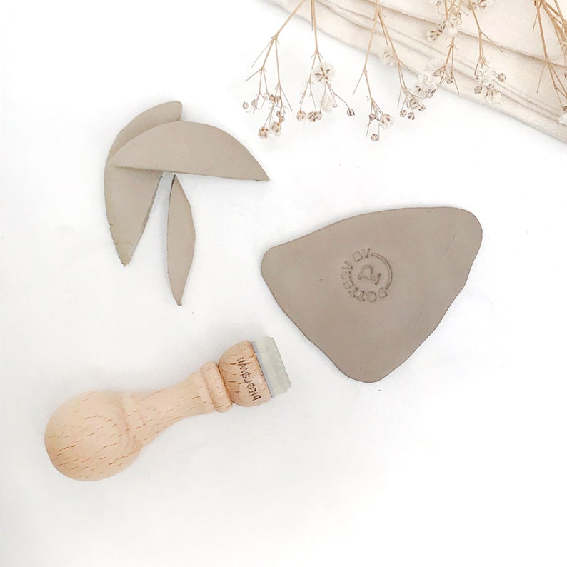 Puede incluir: Un sello de madera con el texto "Dierkswil" en &eacute;l. El sello est&aacute; al lado de un tri&aacute;ngulo de arcilla marr&oacute;n claro con un dise&ntilde;o circular que dice "Made by Pottery".
