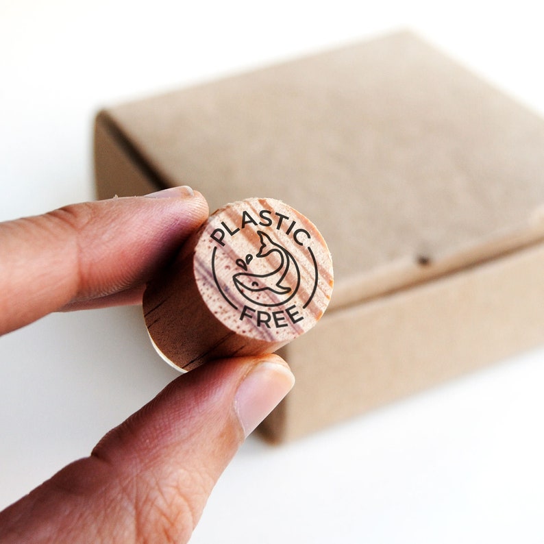 This photo showcases the mini rubber stamp in the palm of a hand, demonstrating its compact size and easy handling. The stamp features a cute whale design and the words "plastic free" around it, making it ideal for stamping eco-friendly packaging materials. This visual emphasizes the practicality and convenience of using this stamp for sustainable shipping.