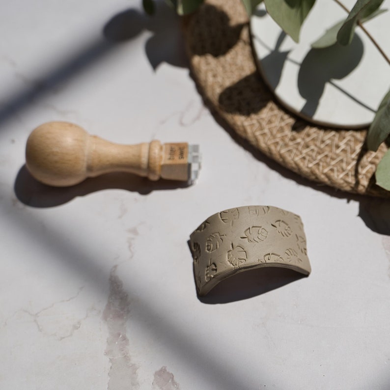 May include: A wooden stamp with a leaf design and a brown clay stamp with a leaf pattern. The clay stamp is curved and has a textured surface.
