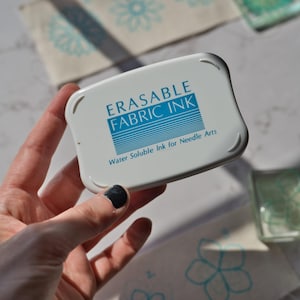 Puede incluir: Un recipiente de plástico blanco con letras azules que dicen "Erasable Fabric Ink" y "Water Soluble Ink for Needle Arts".