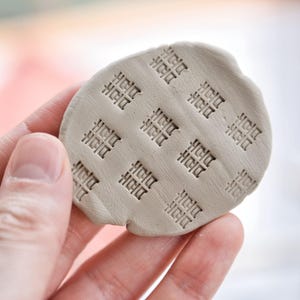 May include: A small, round piece of light gray clay with a grid pattern of Chinese characters pressed into the surface. The clay is held in a hand, with the background slightly blurred.