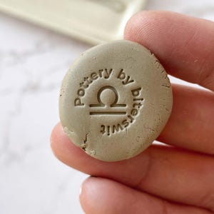 May include: A round, light brown clay stamp with the text "Pottery by Bittersweet" and a Libra zodiac symbol.
