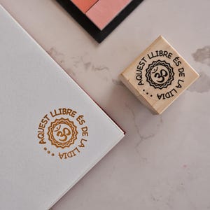 May include: A white notebook with a gold-colored stamp of a sun-like symbol and text in a circular pattern. A wooden stamp with the same design sits nearby. The text reads "Aquest llibre és de la Lidia".