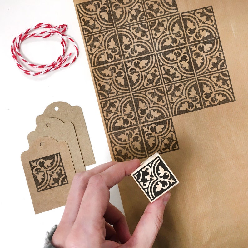 Puede incluir: Una hoja de papel marr&oacute;n con un patr&oacute;n repetido de dise&ntilde;os florales negros. Un sello de madera con un dise&ntilde;o floral negro se est&aacute; utilizando para crear el patr&oacute;n en el papel. Tambi&eacute;n hay varias etiquetas de regalo de papel marr&oacute;n con un dise&ntilde;o floral negro.