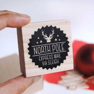 May include: A wooden stamp with a black and white design. The design features a reindeer with antlers, stars, and the text "North Pole Express Mail Via Sleigh".