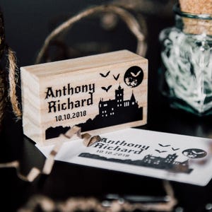 May include: A wooden stamp featuring a black and white illustration of a spooky castle, bats, and a full moon. The text "Anthony Richard 10.10.2018" is printed on the stamp.