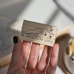 May include: A wooden stamp with a line drawing of a person reading a book. The text on the stamp reads "Ex Libris Laura López".