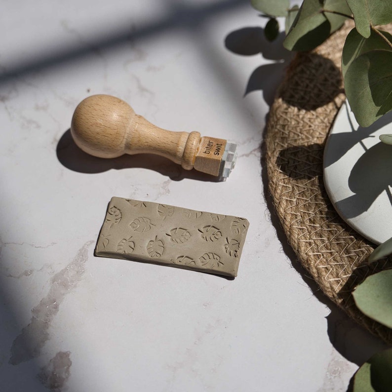 May include: A wooden stamp with the text "biter swit" on it, used to create a pattern of leaves on a piece of clay.