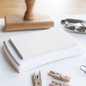 May include: White ink pad with a textured surface, in a white plastic case. The case has a rounded edge.