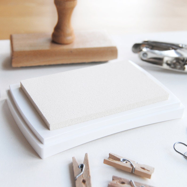 May include: White ink pad with a textured surface, in a white plastic case. The case has a rounded edge.