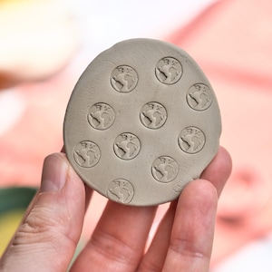 Op de afbeelding: Een ronde, lichtgrijze kleistempel met meerdere reliëf globe-ontwerpen. De stempel wordt in een hand gehouden, met een vage roze achtergrond. De globe-ontwerpen zijn gelijkmatig verdeeld over het oppervlak van de klei.