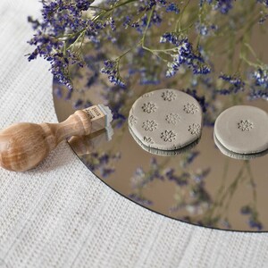 May include: A wooden stamp with the text "Bl&uuml;ten Stempel" on it, next to two gray clay circles. One circle has a pattern of small flowers embossed on it. The clay circles are on a mirrored surface.