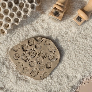 Puede incluir: Una pieza de arcilla beige con un patrón floral impreso, rodeada de arena blanca. Se ven dos sellos con mango de madera y diseños florales, junto con un trozo de coral. La pieza de arcilla mide unos 10 cm de largo.