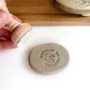 May include: A close-up of a gray clay disc with the text "POTTERY BY MARTHA" and a hand-shaped logo stamped into the clay.