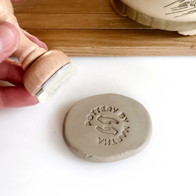 May include: A close-up of a gray clay disc with the text "POTTERY BY MARTHA" and a hand-shaped logo stamped into the clay.