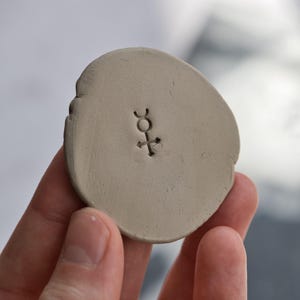 以下が含まれることがあります： スタンプされたシンボルが付いた、小さく、明るいベージュ色の楕円形のセラミック片。シンボルは、矢印が貫通し、湾曲した線で覆われた十字です。この作品は手で持たれています。