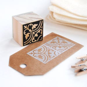May include: A wooden stamp with a black and white floral design. The stamp is used to create a decorative pattern on a brown paper tag.