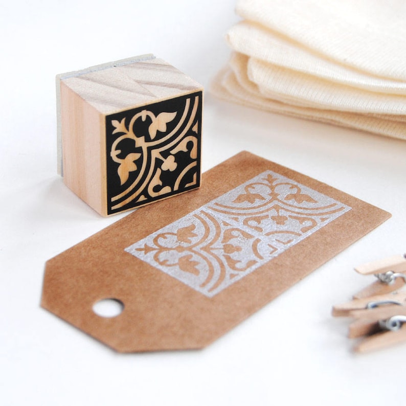 May include: A wooden stamp with a black and white floral design. The stamp is used to create a decorative pattern on a brown paper tag.