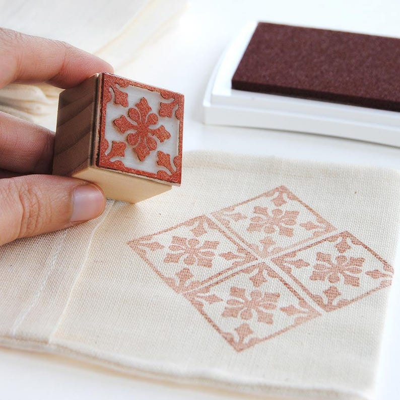 May include: A wooden stamp with a brown and white tile pattern design. The stamp is being used to create a pattern on a piece of natural fabric. The fabric is a light beige color.