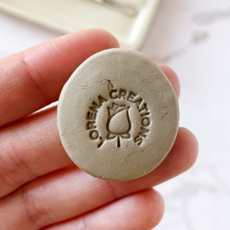 May include: A close-up of a light brown clay stamp with the text "Lorena Creations" and a stylized rose design.