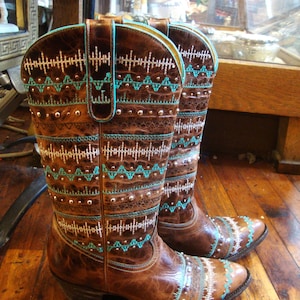 May include: A pair of brown leather cowboy boots with intricate turquoise and white embroidered patterns. The boots feature silver-colored studs and a pointed toe, showcasing a Western-inspired design. The boots are on a wooden floor.