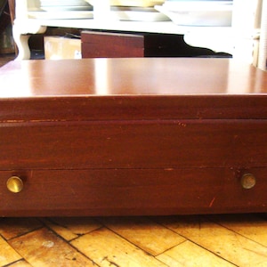 May include: A wooden jewelry box with a rich, dark brown finish. The box has a rectangular shape with a slightly curved top and a single drawer with two brass knobs. The box sits on small feet.
