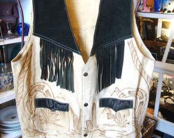 May include: A vintage cream-colored leather vest with black suede accents and fringe. The vest features a desert landscape design with cacti and mountains, two front pockets, and button closures. The vest is hanging on a wooden hanger.