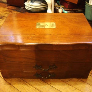 May include: A vintage wooden box with a warm brown finish. It features a brass plate on the top and two drawers with ornate handles. The box is likely used for storage and sits on a wooden floor.