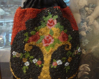 Puede incluir: Un bolso negro de cuentas con un diseño floral en tonos rojos, verdes y amarillos. El bolso tiene un cierre de cordón y un ribete de flecos.