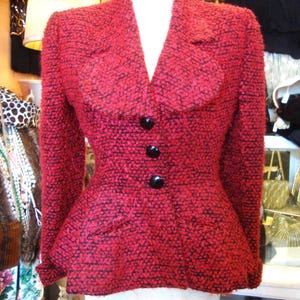 Op de afbeelding: Rode getextureerde blazer met zwarte accenten en drie grote zwarte knopen. De jas heeft een getailleerde pasvorm en een klassieke kraag, gepresenteerd op een mannequin. De blazer is in vintage stijl.