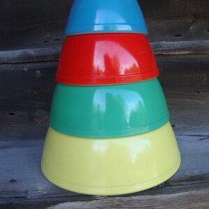 Pode incluir: Um conjunto de quatro taças de mistura empilhadas por ordem decrescente de tamanho. A taça superior é azul brilhante, seguida por uma vermelha, uma verde e uma taça grande amarela na base. Cada taça tem um acabamento liso e brilhante.