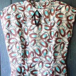 Può includere: Una blusa bianca con un motivo astratto marrone e turchese. La blusa ha un colletto e un fiocco.