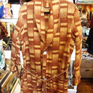 May include: A long-sleeved robe featuring a brown and tan geometric block pattern. The garment has a shawl collar, two front pockets, and a braided cord tie at the waist. It is secured with two white buttons down the front and has braided trim.