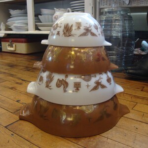 May include: A set of four nesting mixing bowls with a vintage design. The bowls are white with brown and gold accents. The bowls feature a repeating pattern of stylized birds, flowers, and geometric shapes. The bowls are stacked on top of each other, with the largest bowl at the bottom and the smallest bowl at the top.