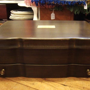 May include: A dark brown wooden box with a rectangular form and a gently curved top. The box features two drawers with brass knobs. The wood grain is visible, and the box appears to be well-crafted.