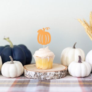 May include: A fall-themed table setting with white pumpkins, a blue pumpkin, a cupcake with an orange pumpkin topper, and a vase with wheat stalks.