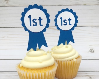 May include: Two yellow cupcakes with white frosting and blue ribbon toppers that say "1st" on them.