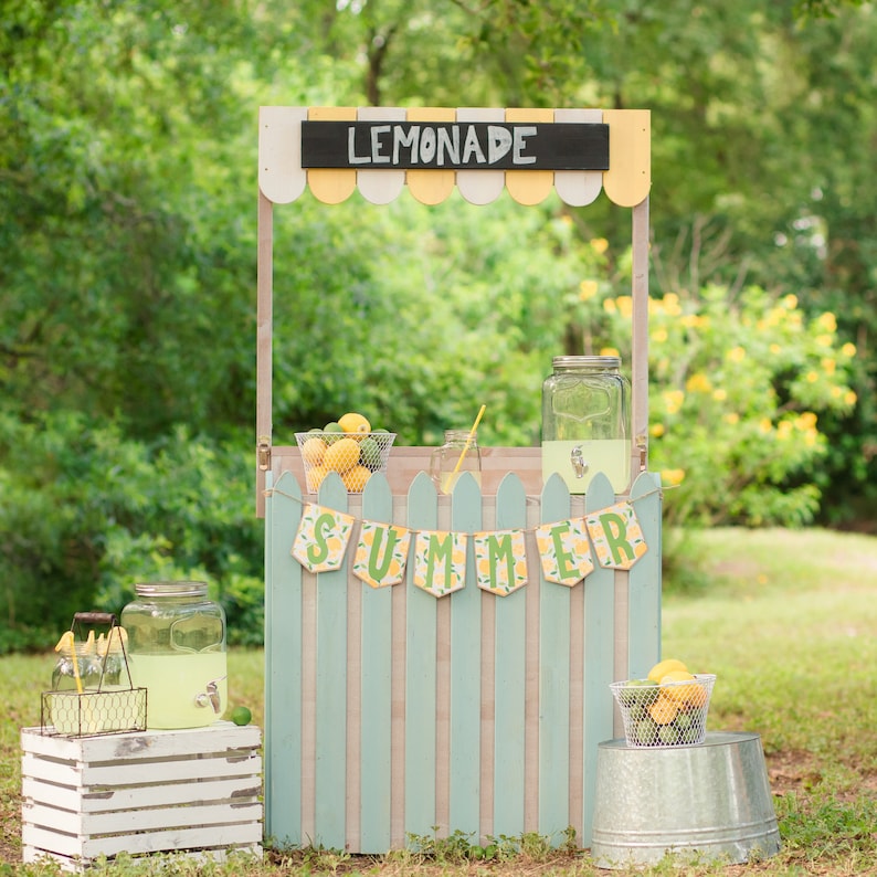 May include: A wooden lemonade stand painted in light blue and white with a yellow awning. The stand has a sign that says "Lemonade" and a banner that says "Summer". There are lemons in a basket and a glass pitcher of lemonade.