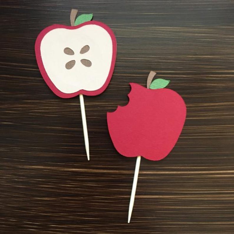 May include: Two red apple cupcake toppers, one whole and one with a bite taken out. The apple toppers are made of felt and have a green stem and leaf.