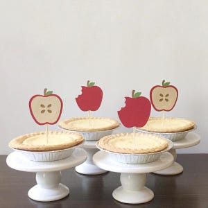 May include: Four mini apple pies topped with red apple and apple slice toppers on white cake stands.