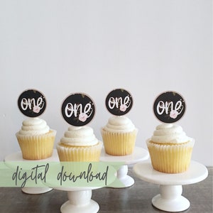 May include: Four cupcakes with black and white floral themed "one" cupcake toppers. The cupcakes are on white cake stands.