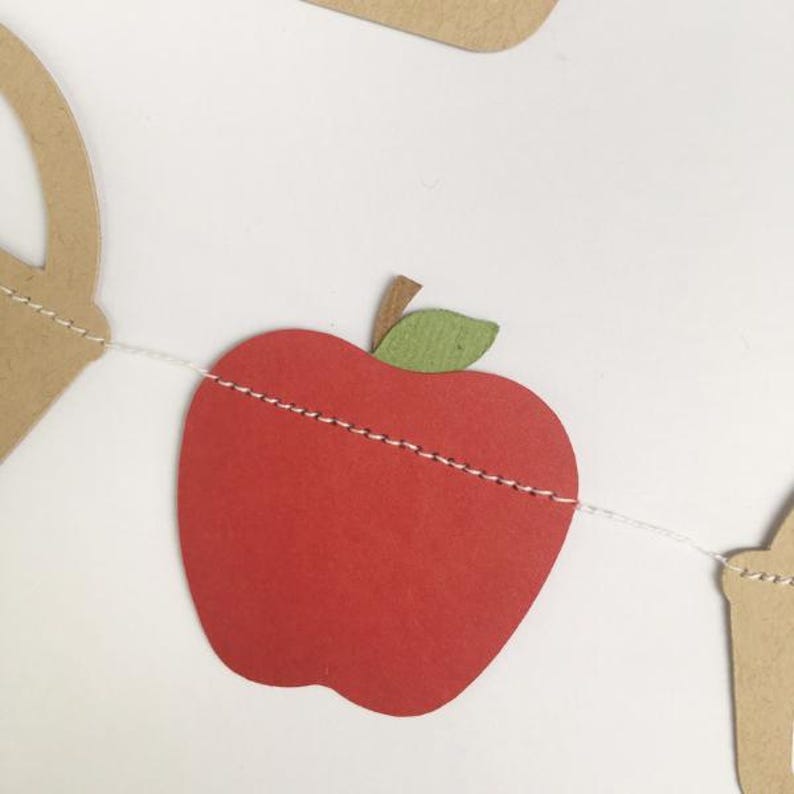 Peut inclure: Une pomme en papier rouge avec une feuille verte, enfil&eacute;e sur une ficelle blanche, fait partie d'une guirlande en papier. La pomme est une forme simple avec une ligne cousue au milieu.