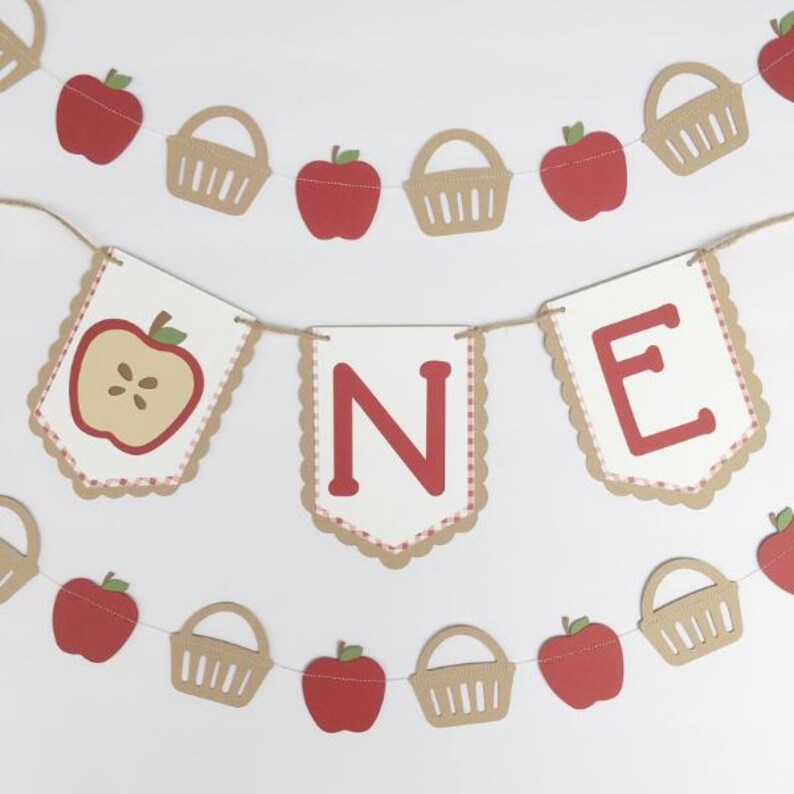 Peut inclure: Une banni&egrave;re sur le th&egrave;me de la pomme rouge et blanche avec le mot "ONE" en lettres rouges. La banni&egrave;re est d&eacute;cor&eacute;e de pommes rouges, de paniers bruns et d'un motif vichy blanc et rouge.