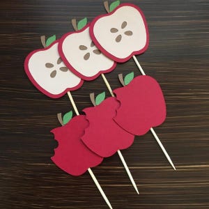 May include: Five red apple cupcake toppers with green stems and leaves. The apples are cut in half and some have a bite taken out of them.