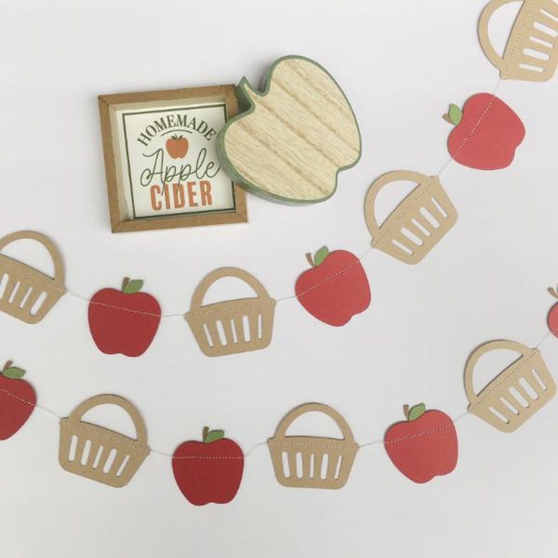 Peut inclure: Une guirlande d'appels rouges et de paniers bruns avec le texte "Homemade Apple Cider" sur une pancarte en bois.