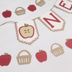 Peut inclure: Une banni&egrave;re sur le th&egrave;me de la pomme rouge et blanche avec le mot "ONE" en lettres rouges. La banni&egrave;re est d&eacute;cor&eacute;e de pommes rouges, de paniers bruns et d'un motif vichy rouge et blanc.