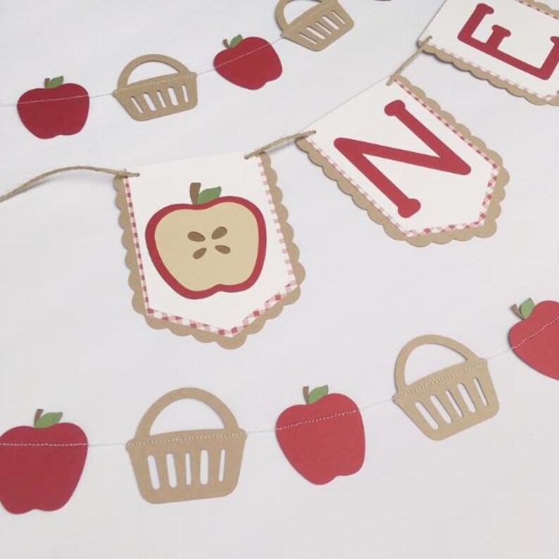 Peut inclure: Une banni&egrave;re sur le th&egrave;me de la pomme rouge et blanche avec le mot "ONE" en lettres rouges. La banni&egrave;re est d&eacute;cor&eacute;e de pommes rouges, de paniers bruns et d'un motif vichy rouge et blanc.